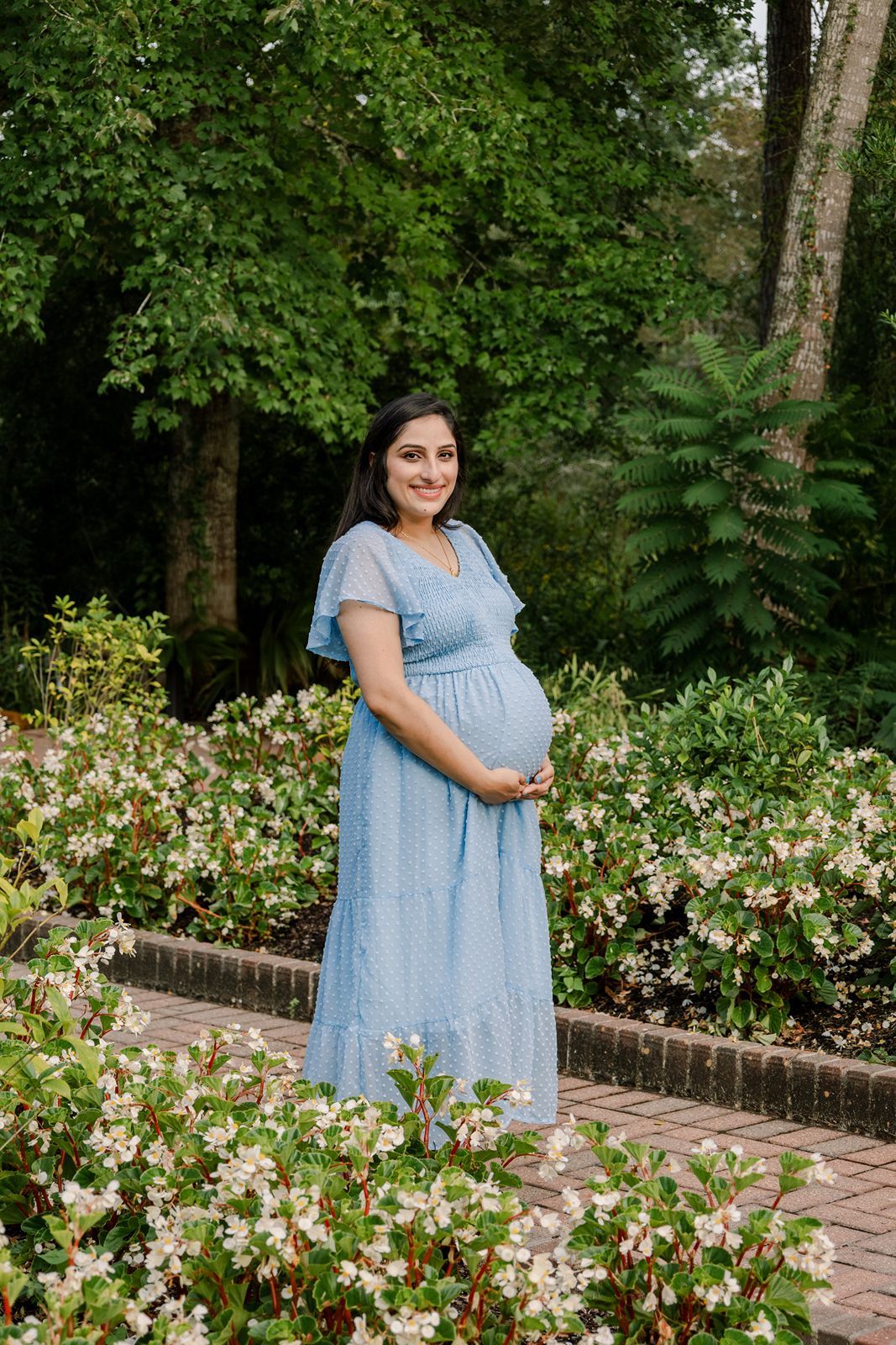 A woman in a light blue dress poses in a garden path surrounded by blooming white flowers and greenery.