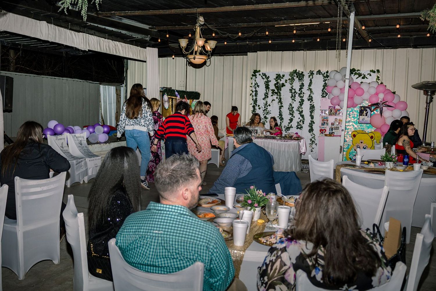 Indoor party venue with guests seated at round tables decorated for a celebration with floral backdrop and string lights.