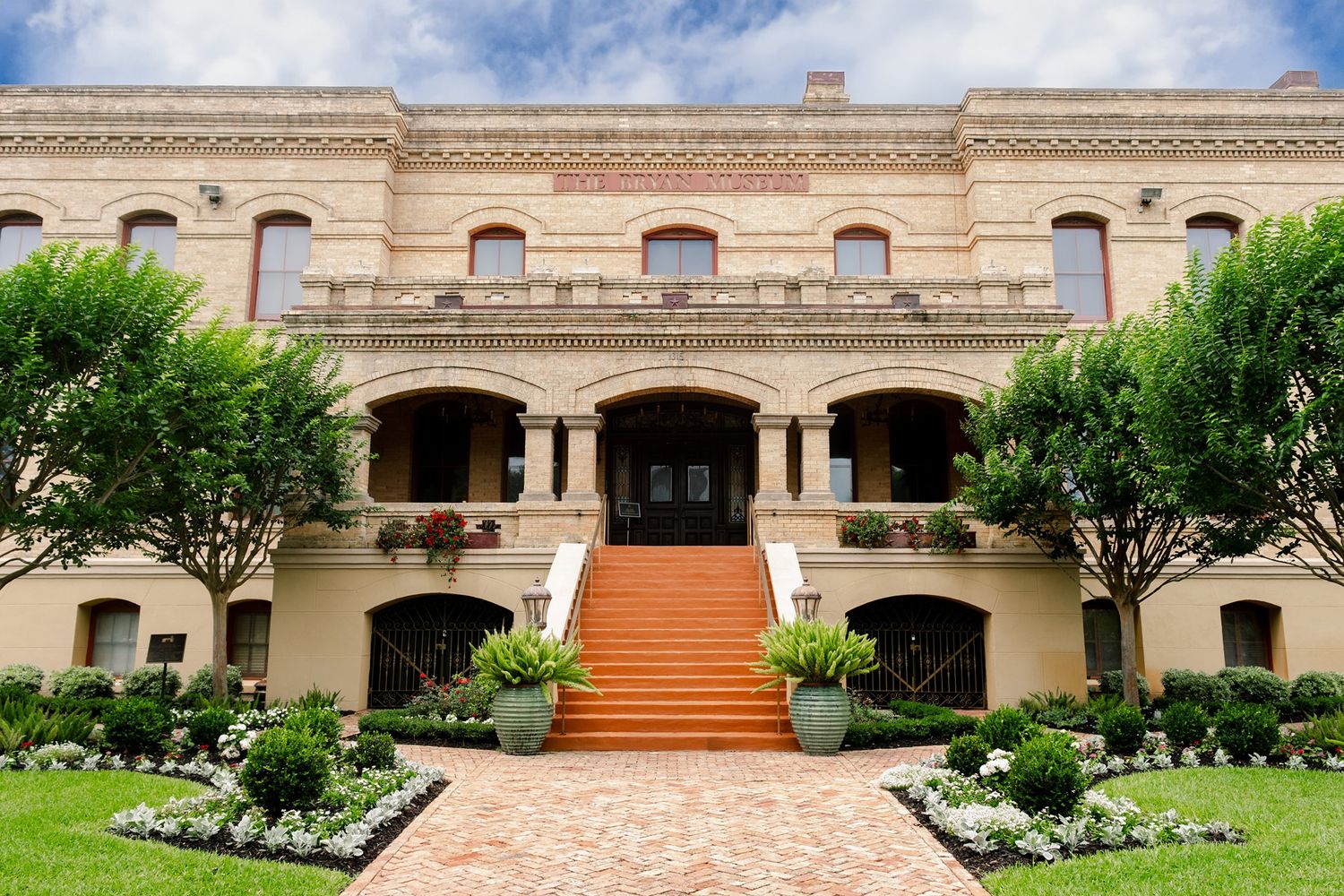 Grand historic beige brick mansion with red staircase, arched windows, manicured gardens, and lush green trees under blue sky.