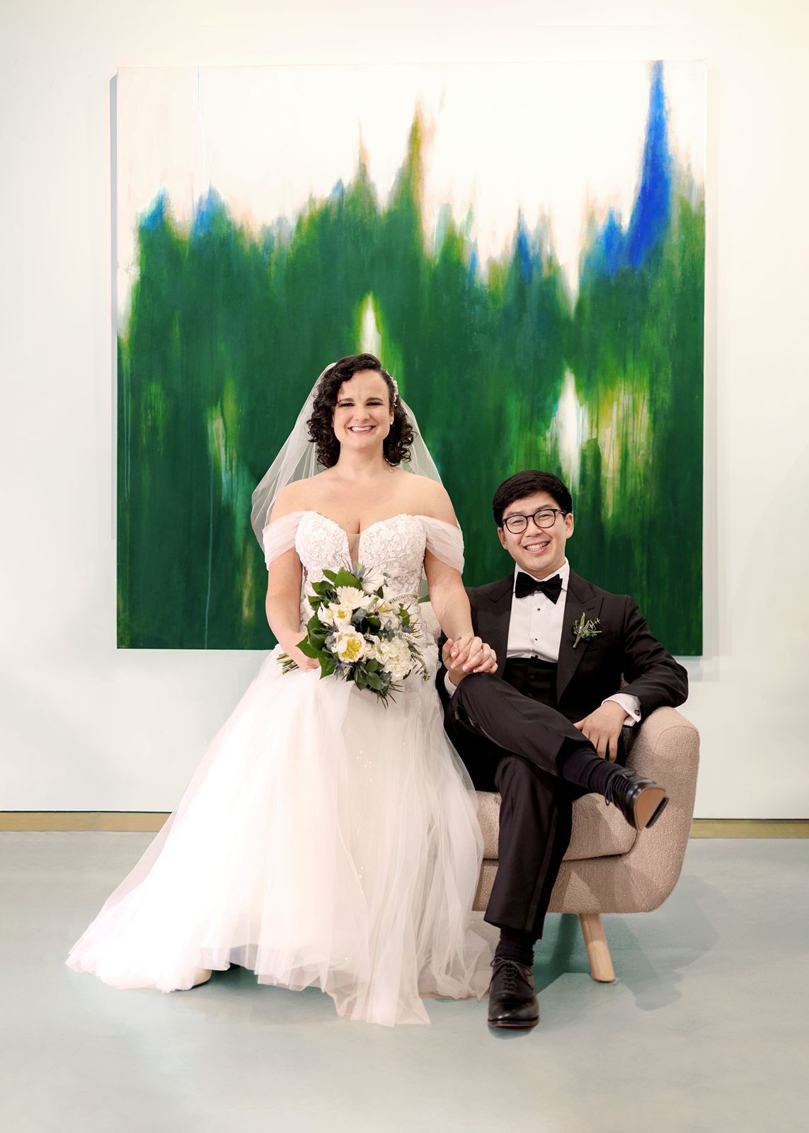 A newlywed couple poses together on a modern white couch with green abstract art in the background.