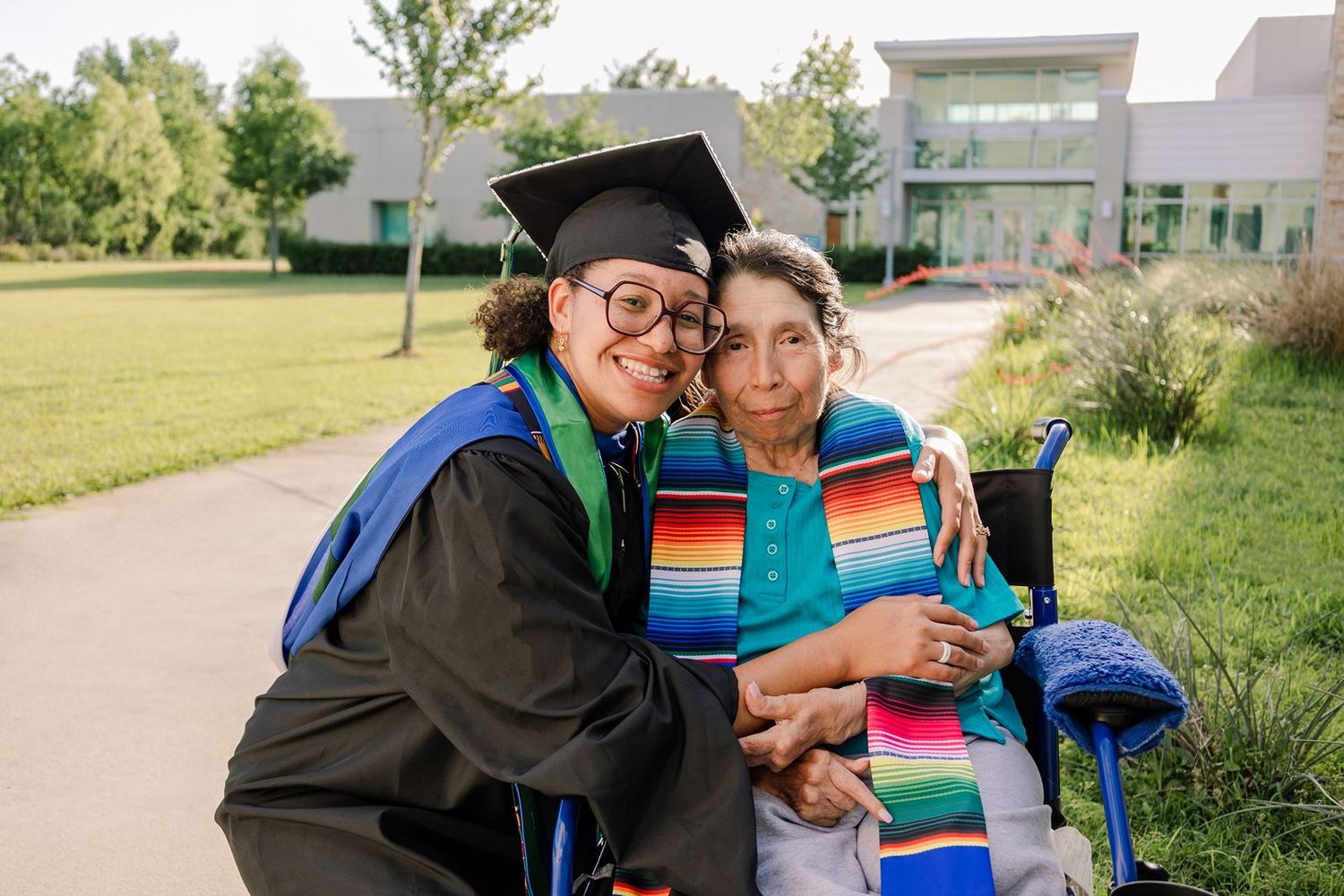 Graduate in cap and gown hugs elderly woman in wheelchair outdoors, both smiling warmly during graduation celebration.