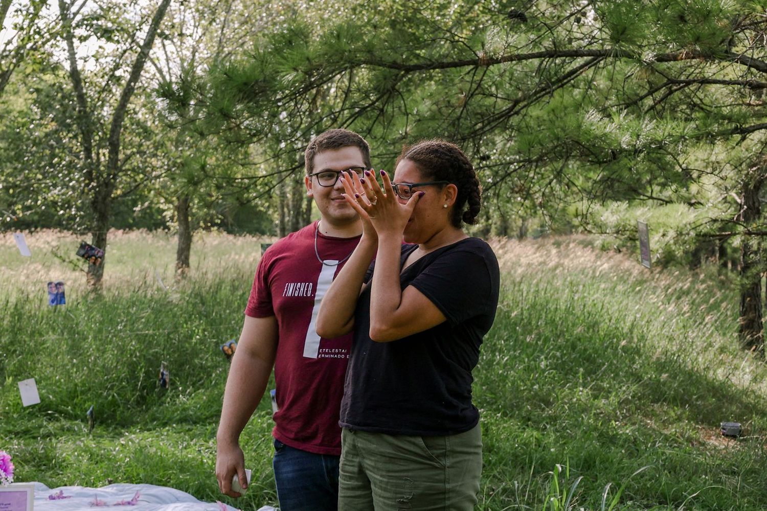 A man and woman stand outdoors near trees, with the woman raising binoculars to her eyes in a green field.