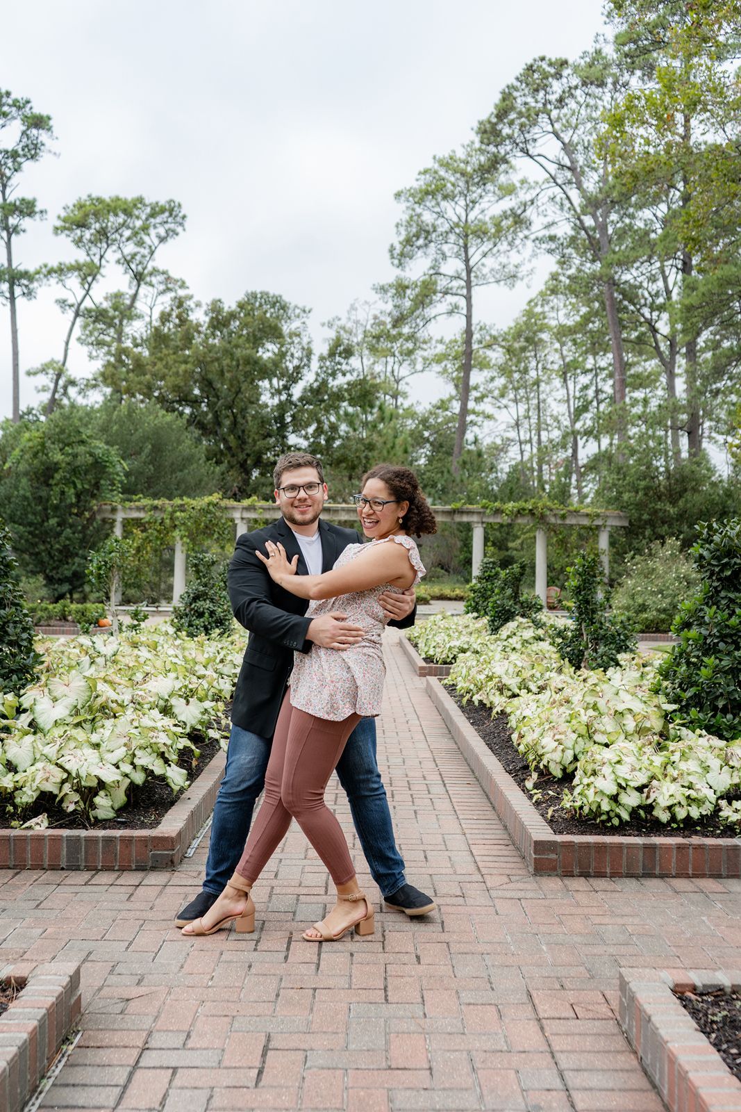 A joyful couple dances together on a brick path in a lush garden with white flowers.