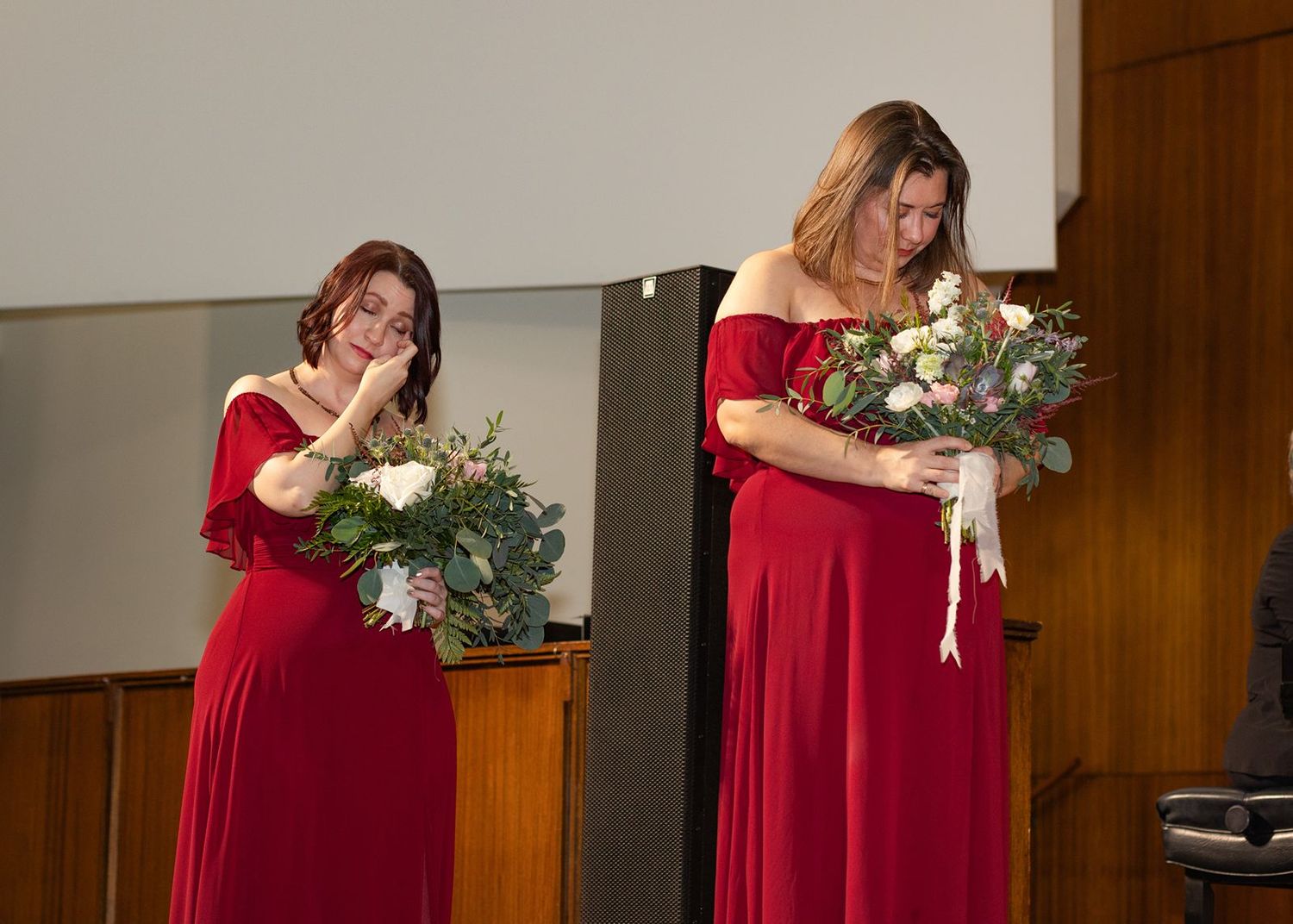 Two bridesmaids in red off-shoulder gowns holding white floral bouquets during wedding ceremony.