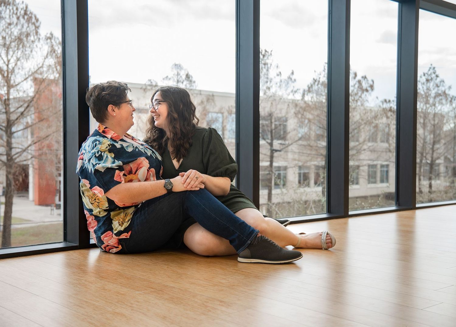 Two people sit on a wooden floor next to large windows in a bright, modern space.