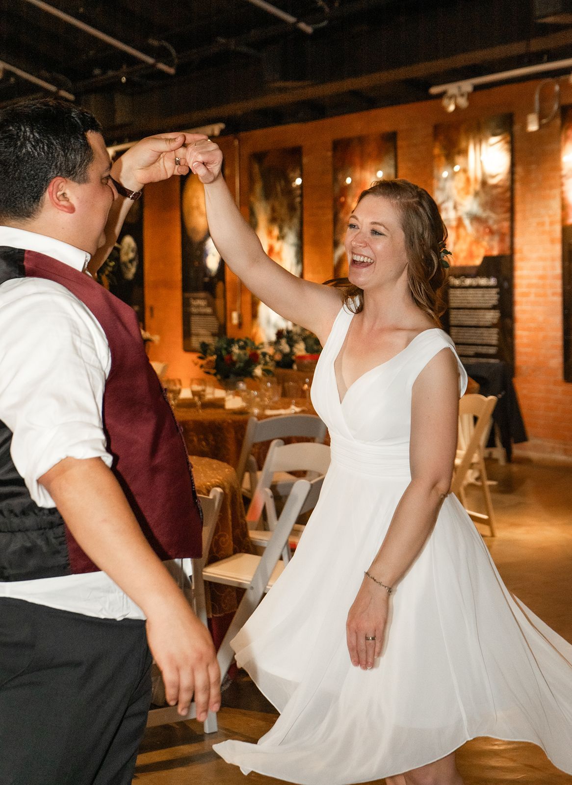 A couple in formal attire dances together in a warmly lit venue with exposed brick walls and pendant lighting.