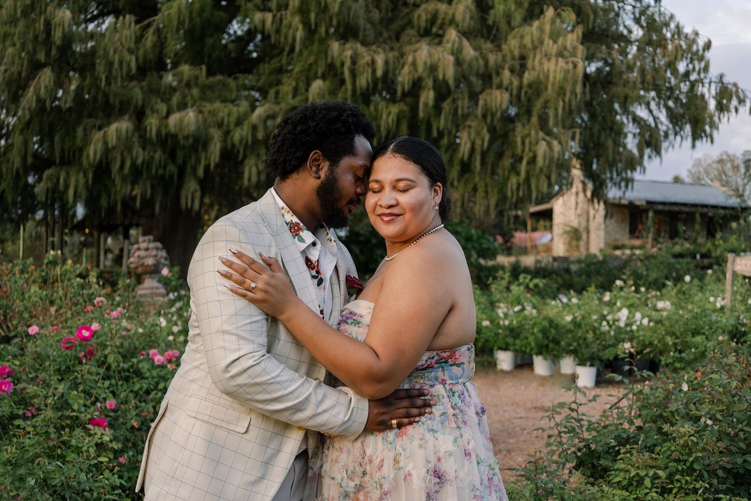 A romantic couple embraces in a garden setting with pink roses and greenery in the background.