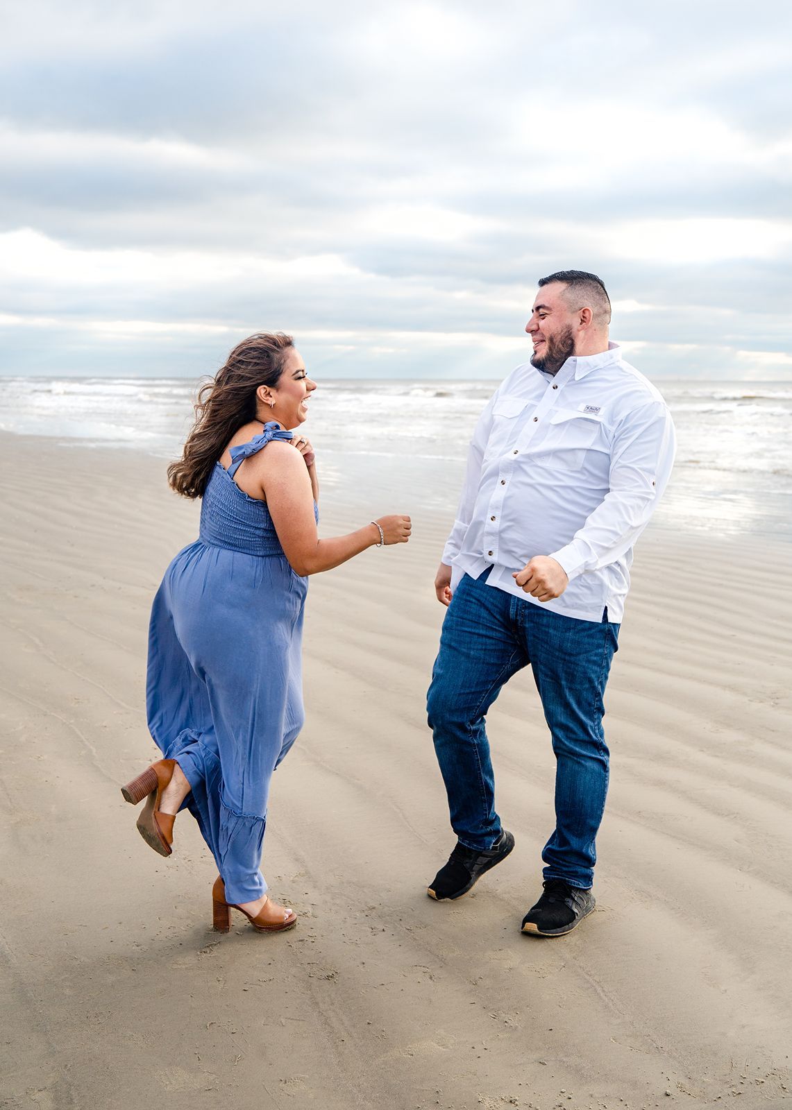 A couple playfully interacts on a cloudy beach, with the woman in a blue dress and the man in a white shirt and jeans.
