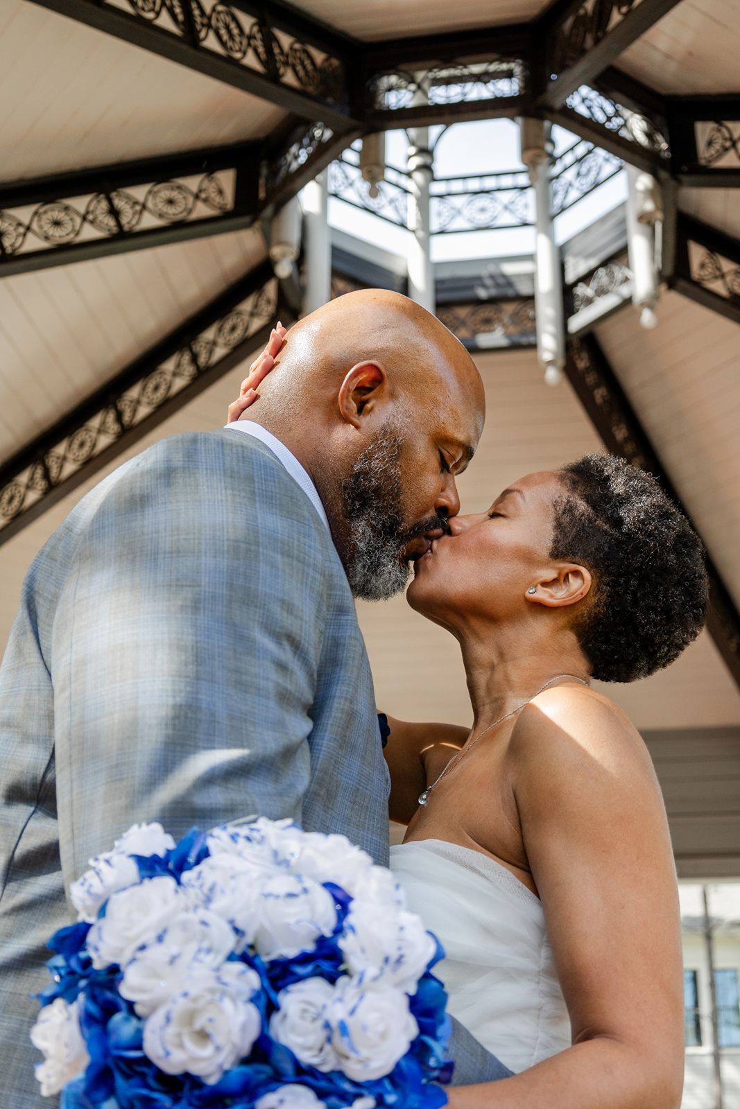 A newlywed couple shares a romantic kiss under a gazebo, bride holding blue and white bouquet.
