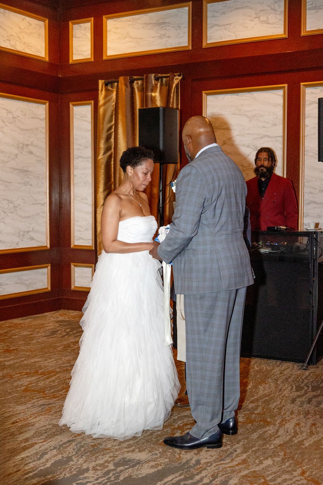Bride in white strapless gown holds hands with groom in gray suit during wedding ceremony with DJ in background.