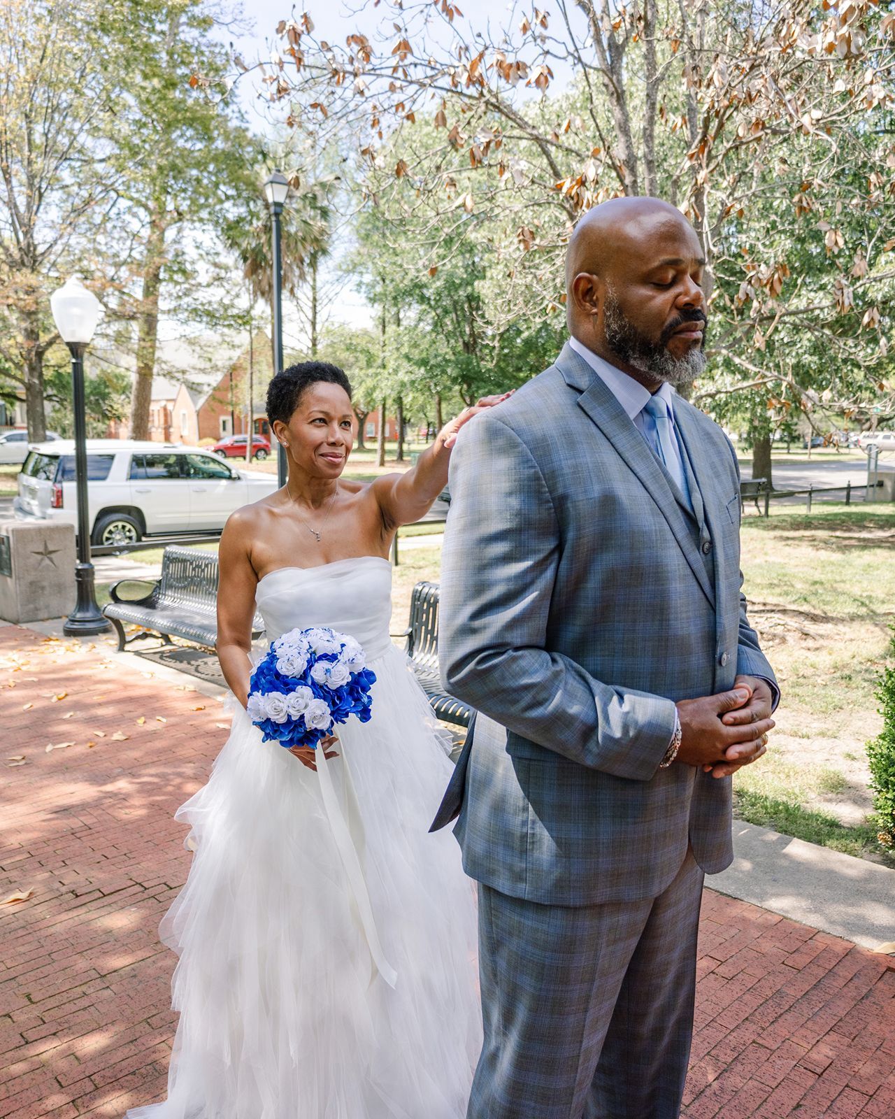Wedding first look moment as bride in white gown taps groom's shoulder holding royal blue bouquet outdoors.