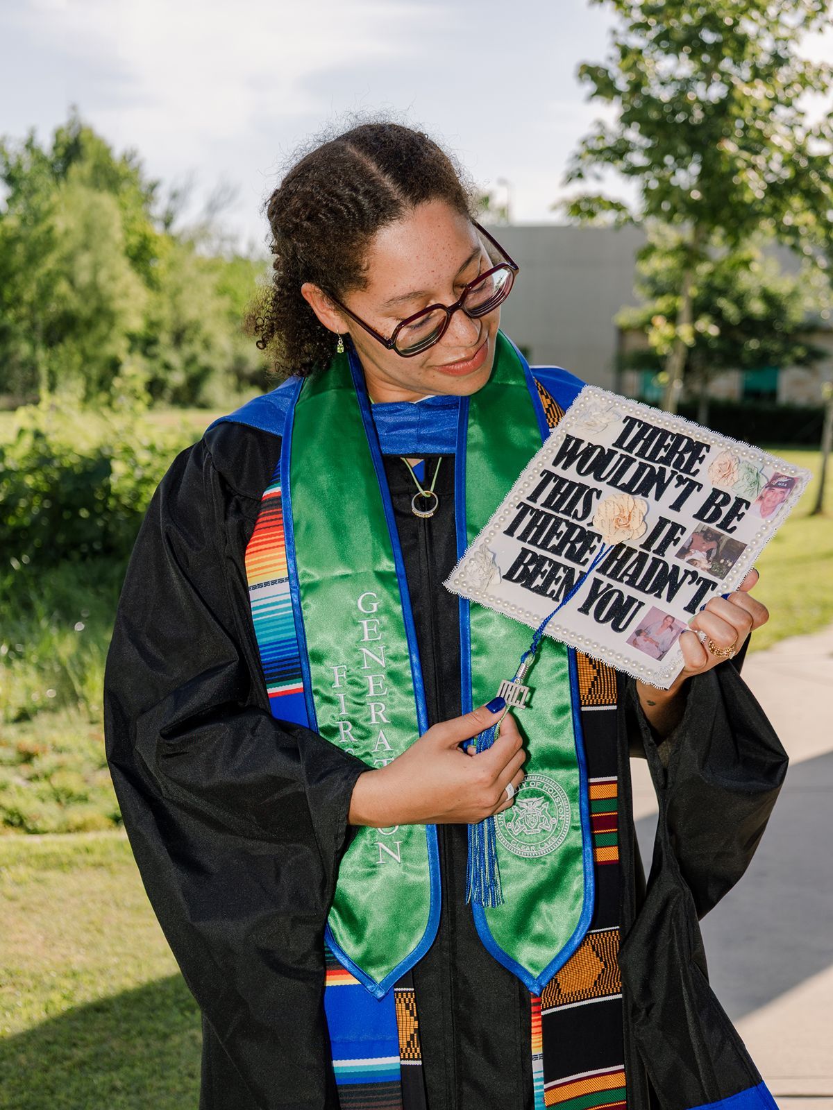 Graduate in black gown with colorful stoles smiling at decorated cap reading There Wouldn't Be This If There Hadn't Been You.