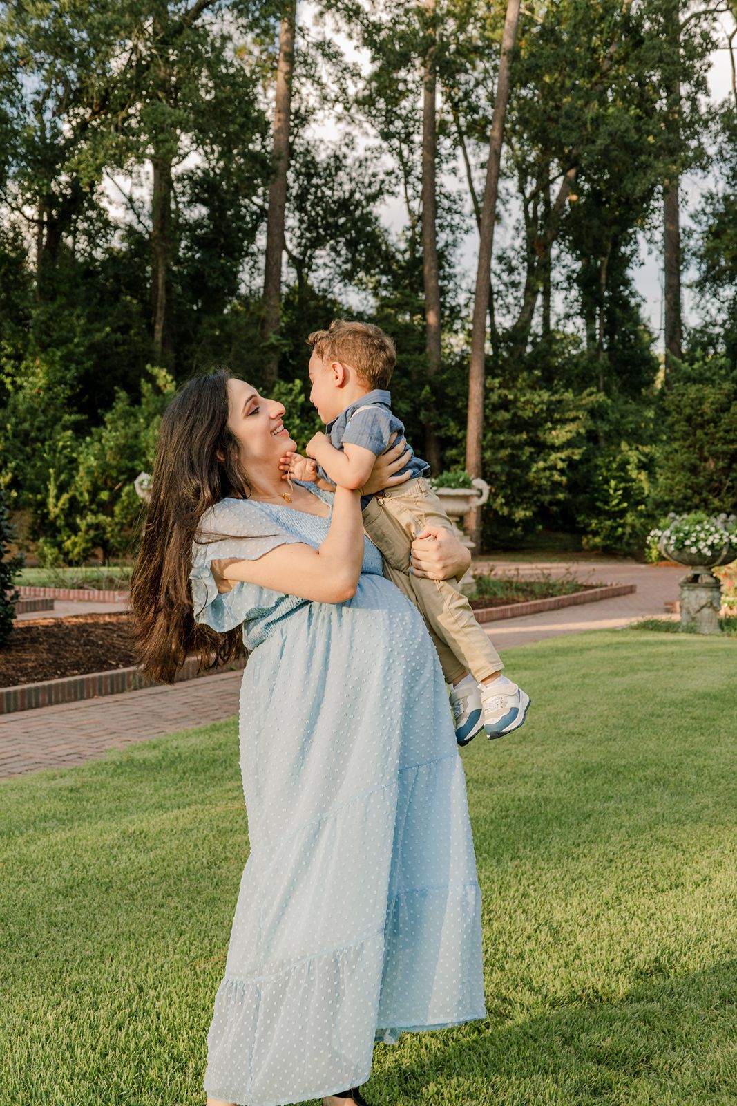 A mother in a blue dress holds and kisses her baby in a park setting with trees and grass in the background.