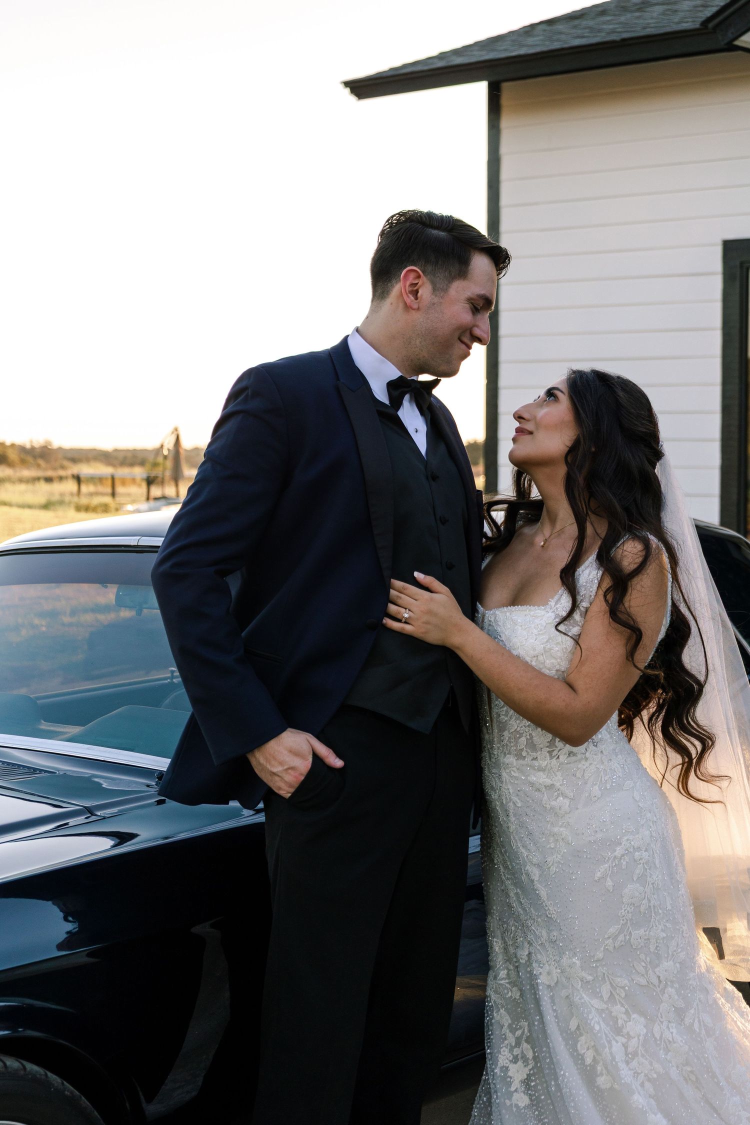 Bride and groom share a romantic moment leaning against a black car, smiling at each other in wedding attire.