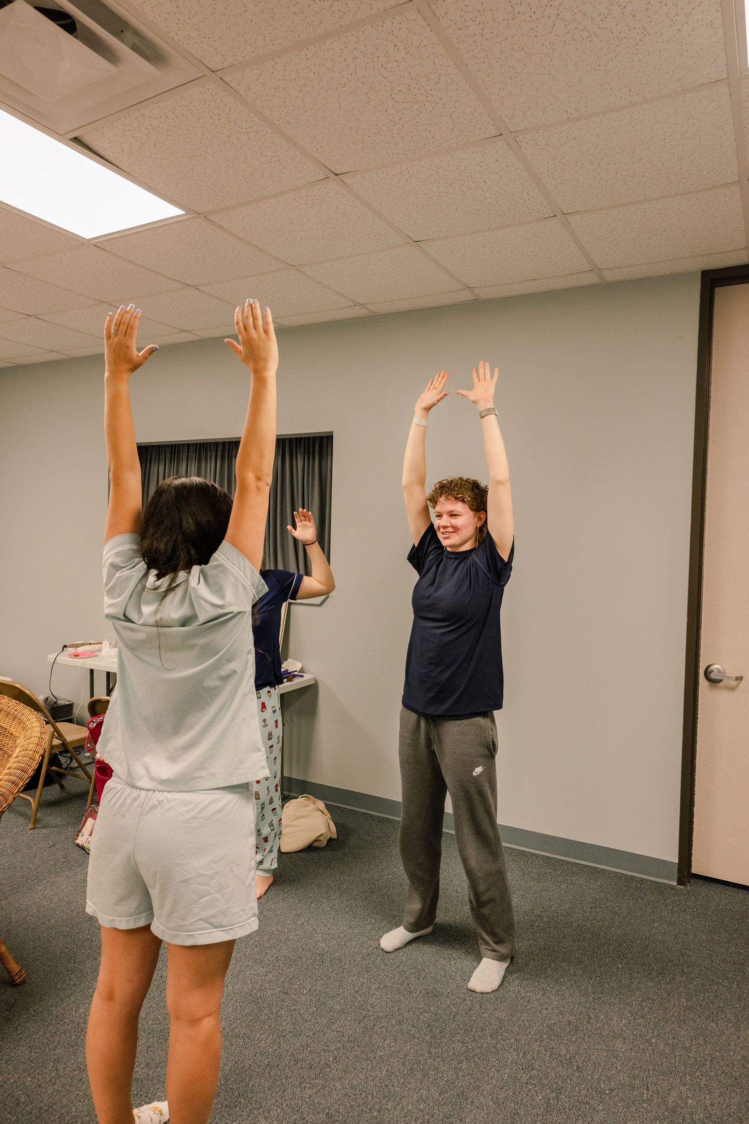 People doing energetic dance moves and stretches in a grey carpeted room.
