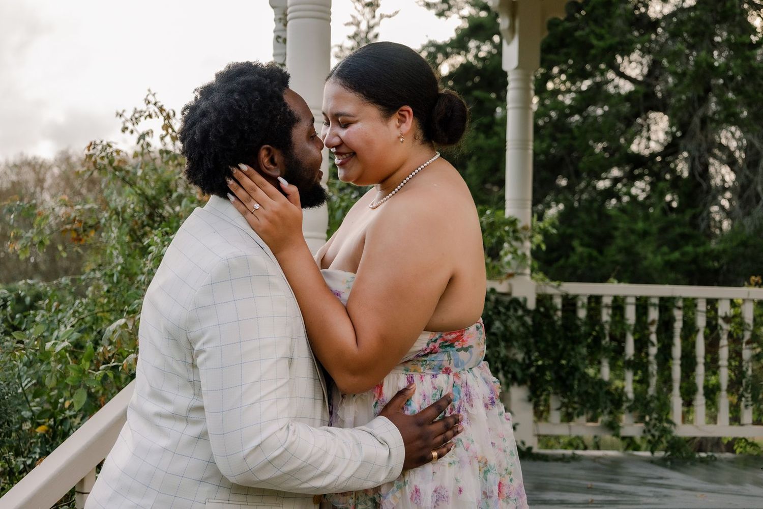 A romantic couple shares a tender kiss on a white porch surrounded by lush greenery and flowers.