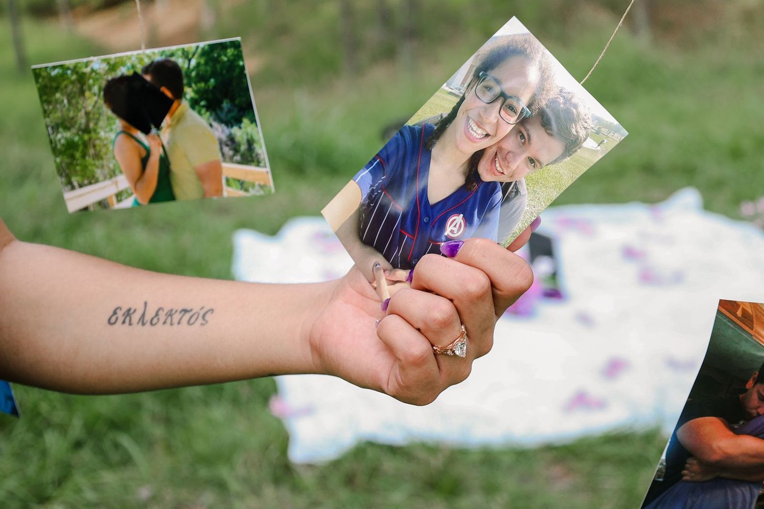 A hand with a wrist tattoo holds a photo of a smiling couple outdoors, with more hanging photos visible in the background.