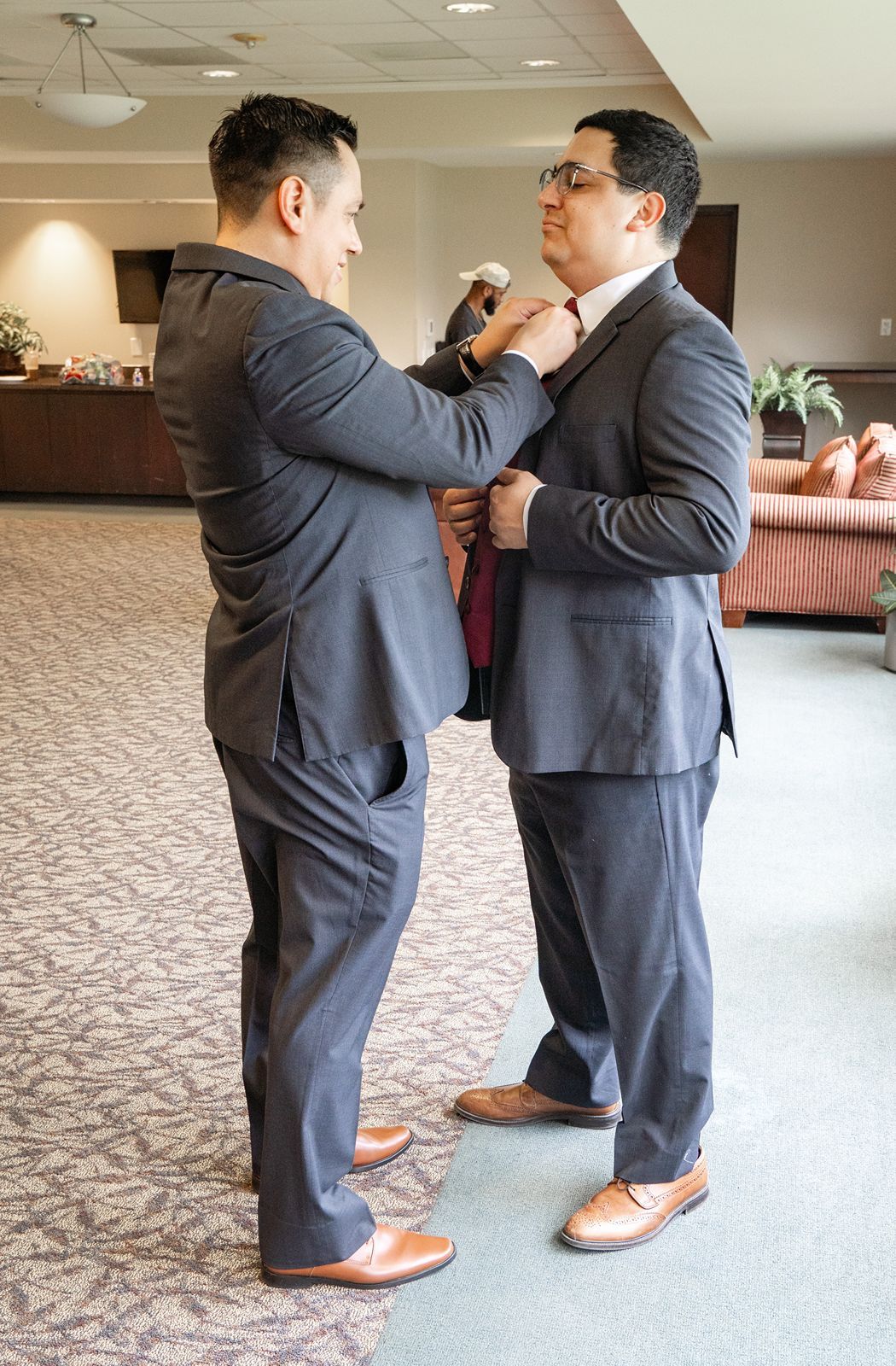 Two men in matching gray suits adjusting a burgundy tie indoors before a wedding ceremony.