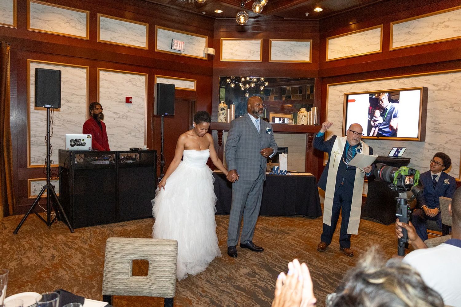Newlywed couple dance at indoor wedding reception with DJ, officiant, and guests celebrating in stylish venue.