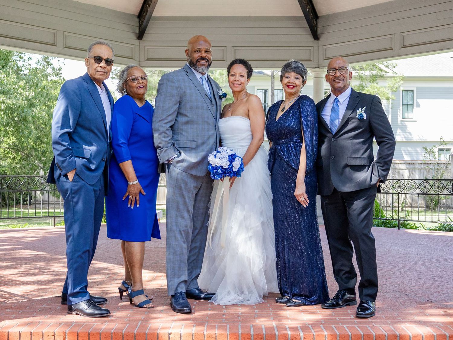 Wedding party posing under gazebo, bride in white gown with blue bouquet, guests in blue formal attire.