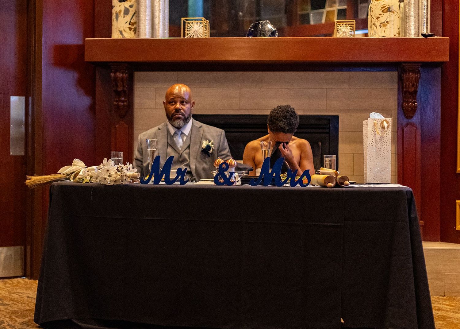 Newlywed couple seated at Mr. and Mrs. sweetheart table with fireplace backdrop at elegant wedding reception.