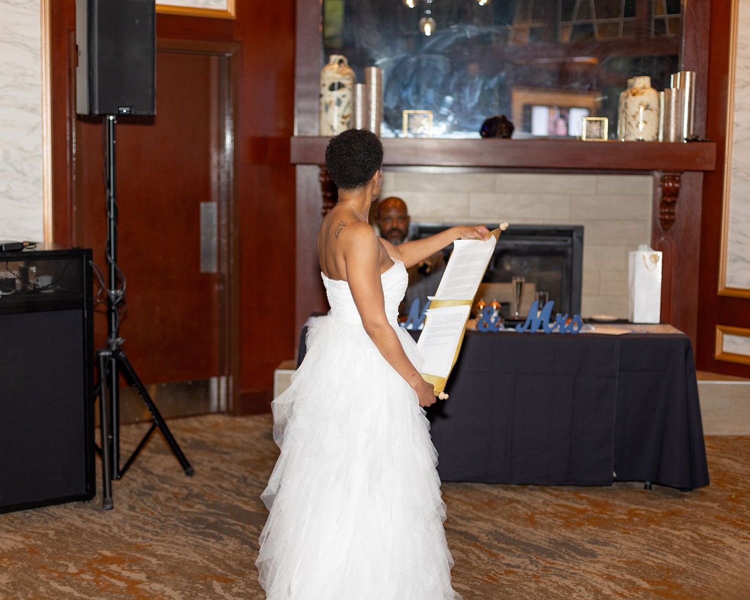 Bride in white ruffled gown reads scroll at indoor wedding ceremony.