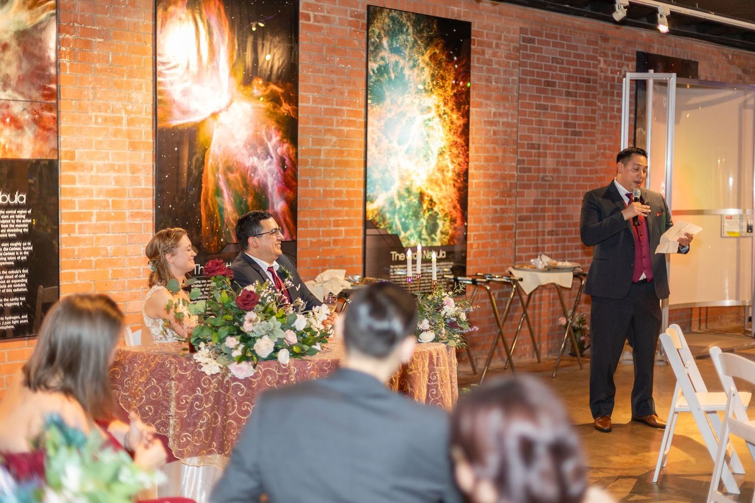 Man giving a toast speech at an elegant wedding reception with brick walls, floral centerpieces, and colorful artwork.