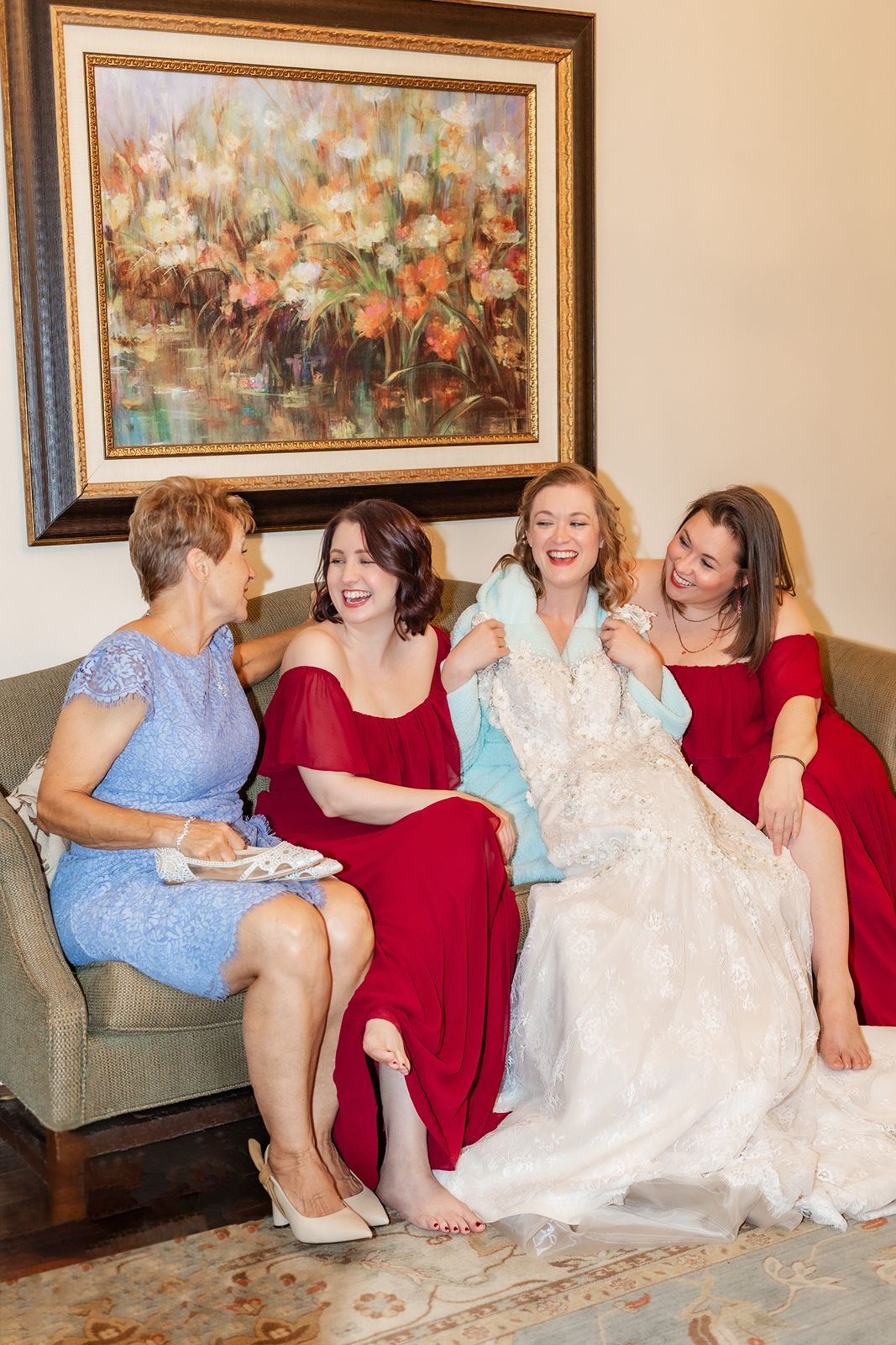 Bride in wedding gown laughs with bridesmaids in red dresses and mother on a couch before the wedding.