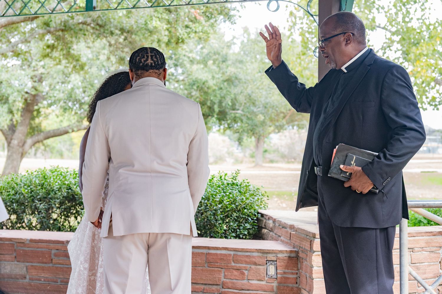 Wedding guests in formal wear standing along a brick wall during an outdoor ceremony.