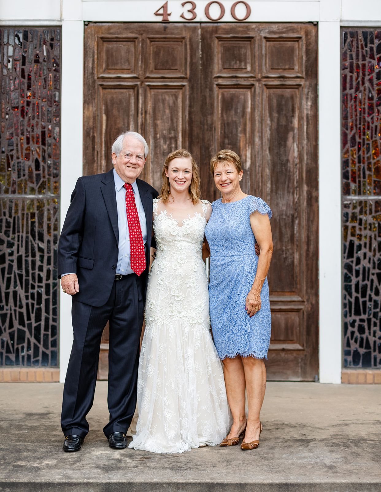 Bride in lace wedding gown poses with parents outside church entrance numbered 4300.
