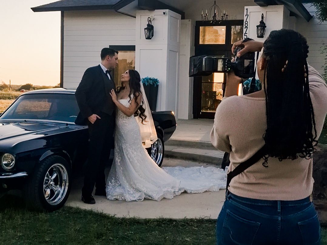 Photographer capturing bride and groom posing beside a classic black car at golden hour outside a white house.