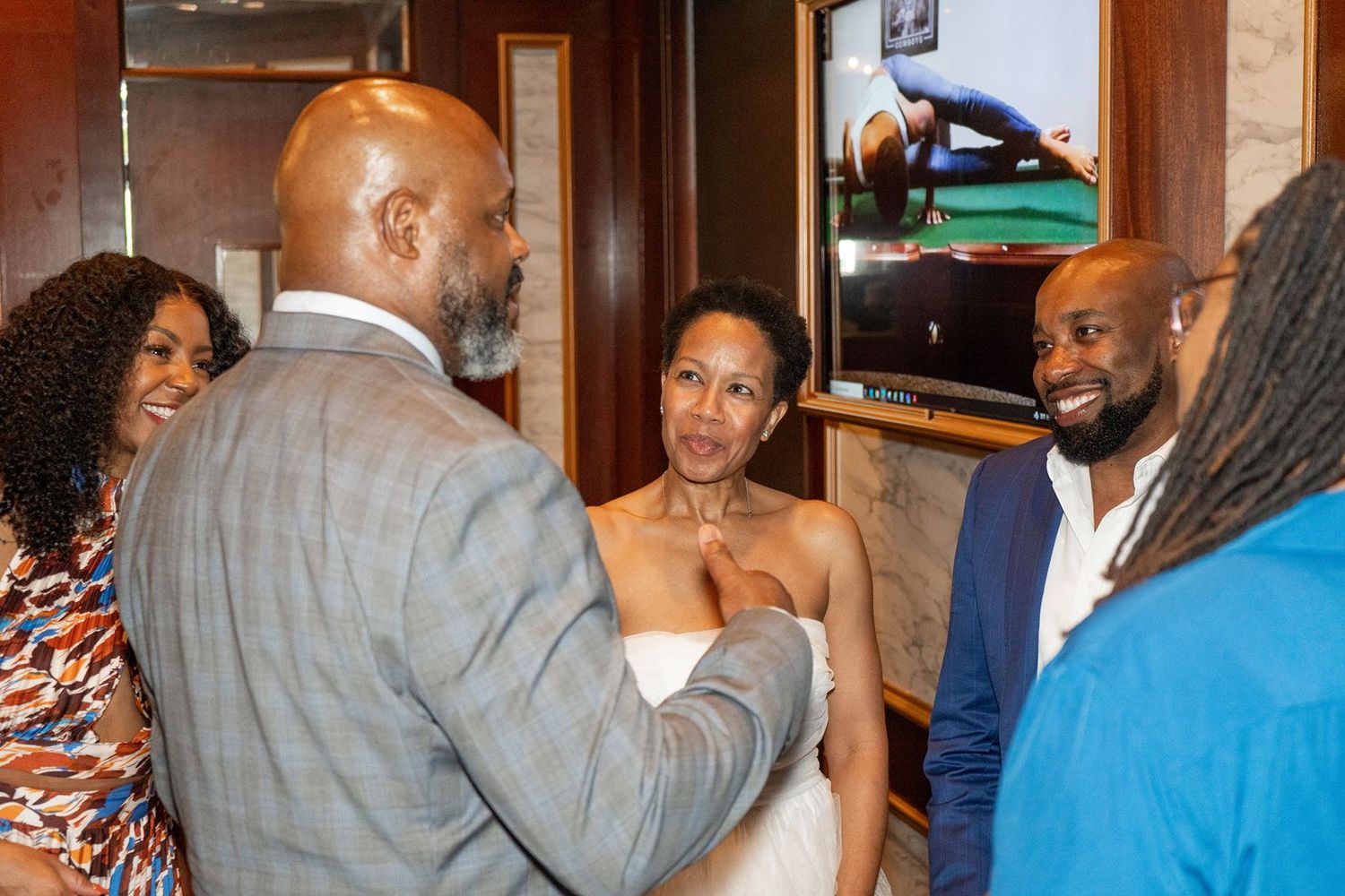Wedding guests in colorful attire congratulate bride in white dress and groom in gray suit at reception.