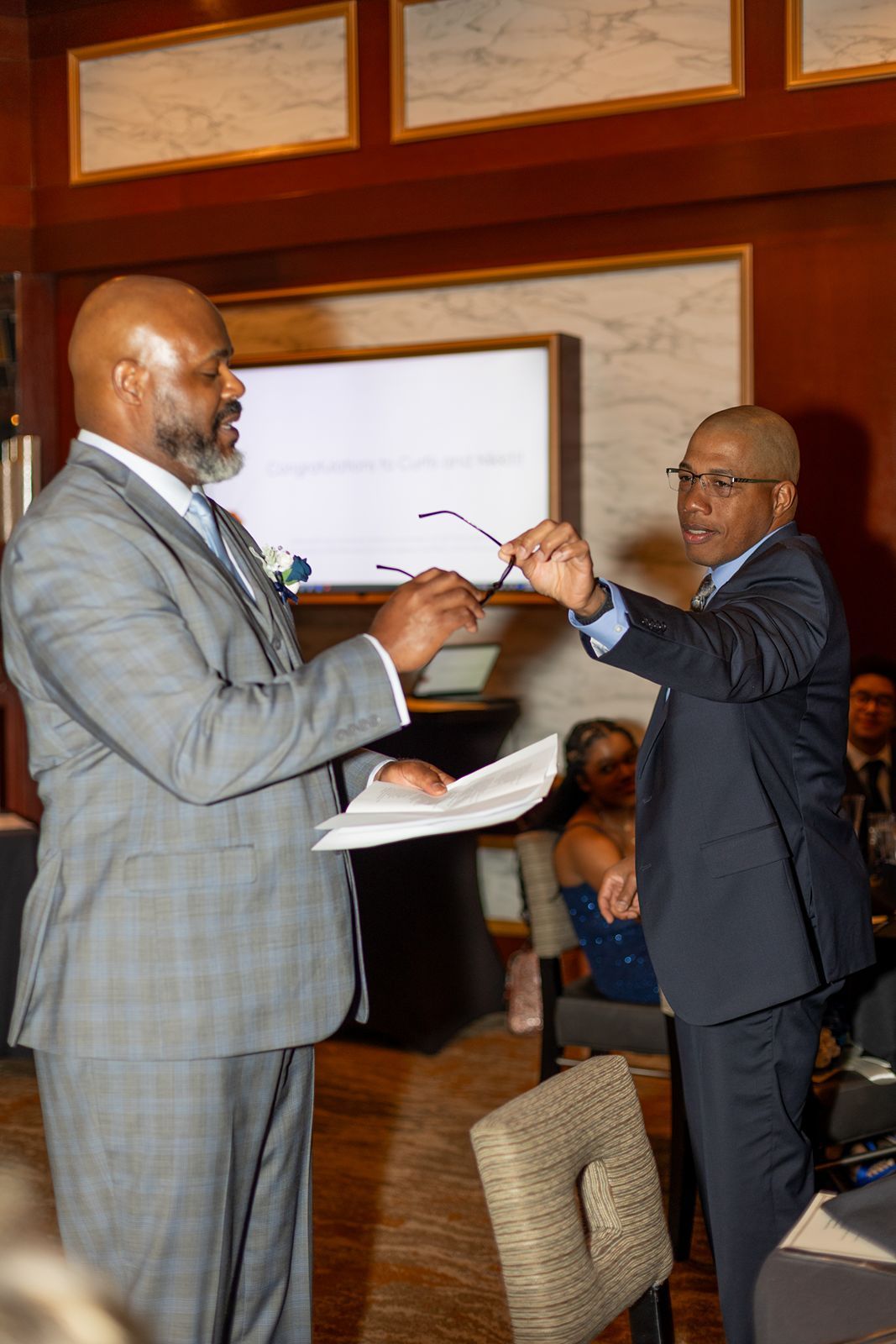 Two men in suits stand facing each other during a formal ceremony, one holding glasses and papers indoors.
