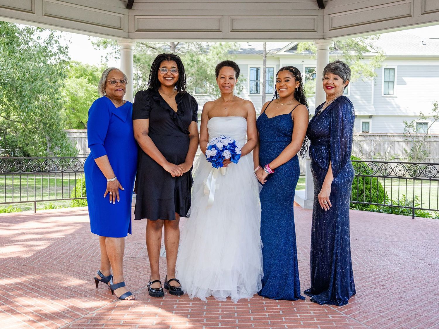 Bride in white gown poses with four women in blue dresses under a gazebo at an outdoor wedding.