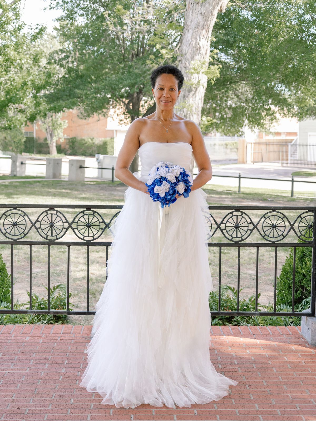 Bride in strapless white tulle gown holding blue and white bouquet outdoors at a wedding.