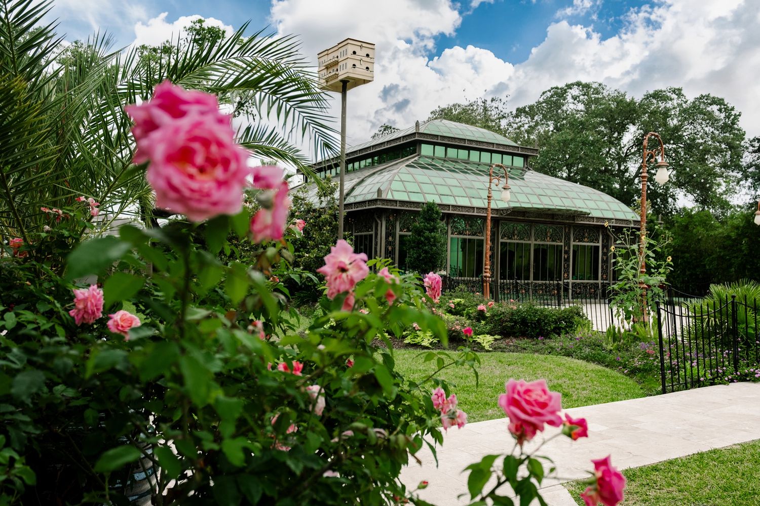 Pink roses bloom in front of a green glass greenhouse pavilion surrounded by lush garden greenery.