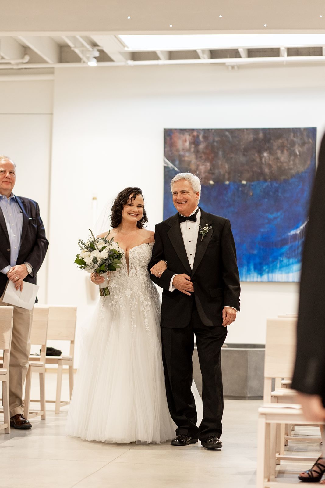 A wedding ceremony processional takes place in a modern white gallery space with blue artwork on display.