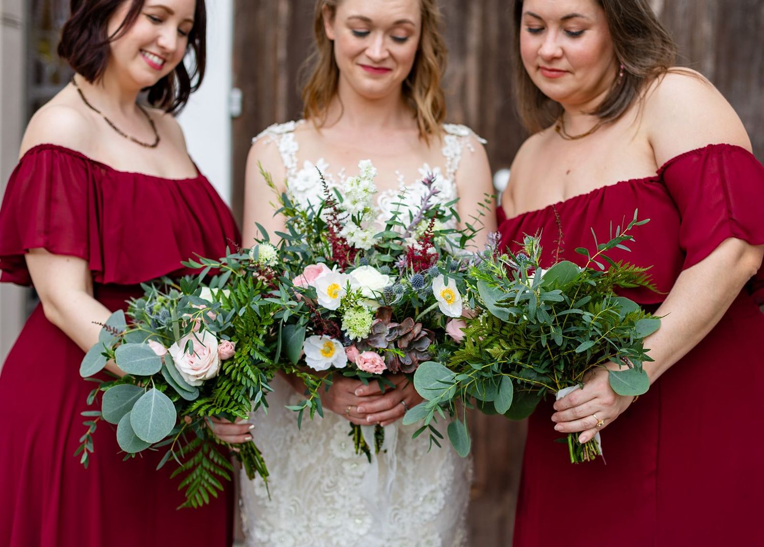 Bride in lace gown holds lush bouquet flanked by bridesmaids in red off-shoulder dresses at rustic barn wedding.
