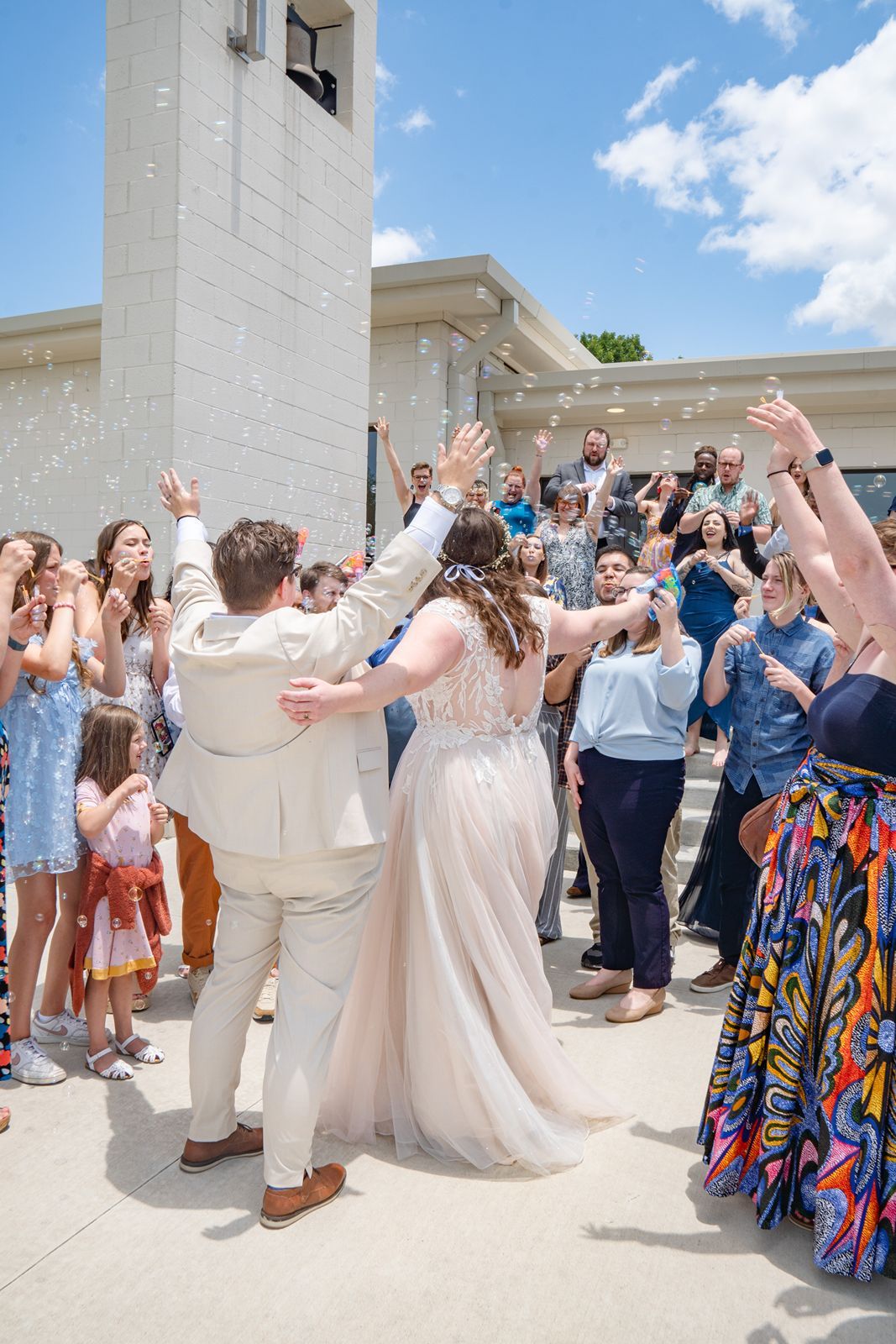Newlyweds exit church surrounded by cheering guests blowing bubbles on sunny wedding day.