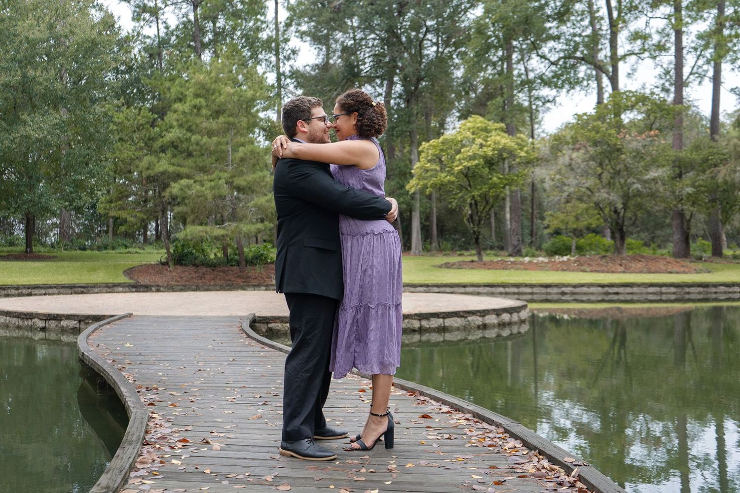 Couple embracing on a wooden dock beside a tranquil pond surrounded by lush green trees.