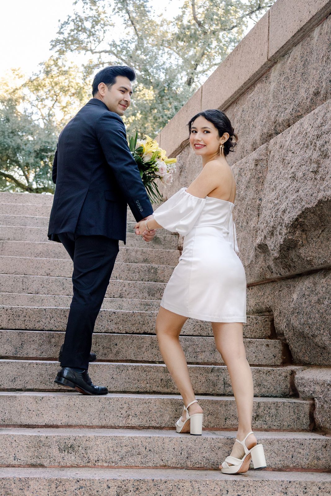 Couple in wedding attire holding hands on stone steps, woman in white mini dress with bouquet, man in black suit.