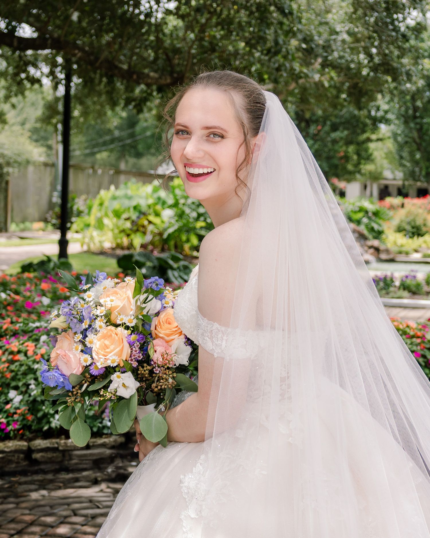 An elegant bridal portrait showcasing a bouquet of peach and purple flowers in a garden setting.