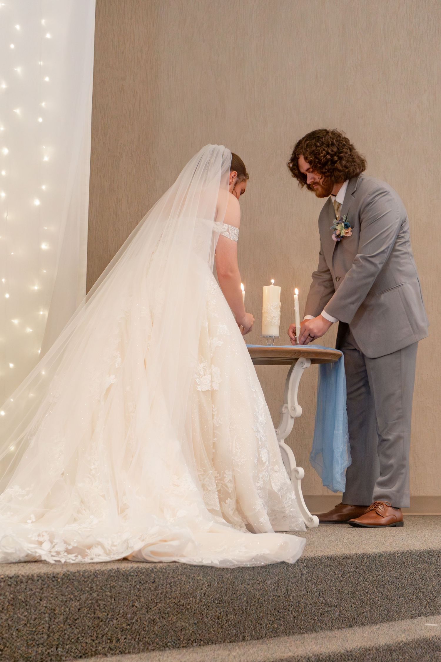 A couple in formal attire share a quiet wedding ceremony moment under a backdrop of twinkling lights.