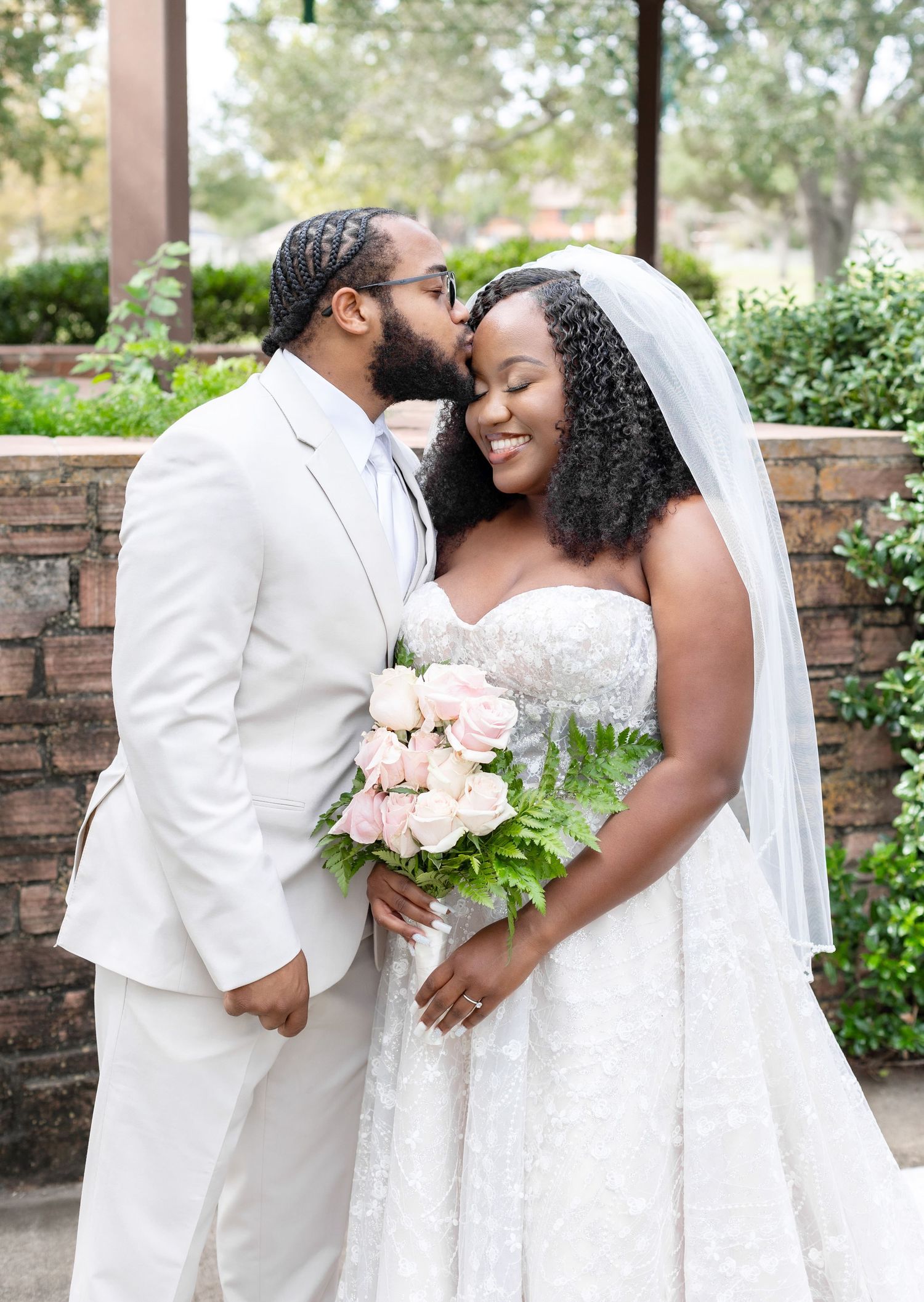 A couple in elegant white wedding attire embraces while holding pink roses in a garden setting.