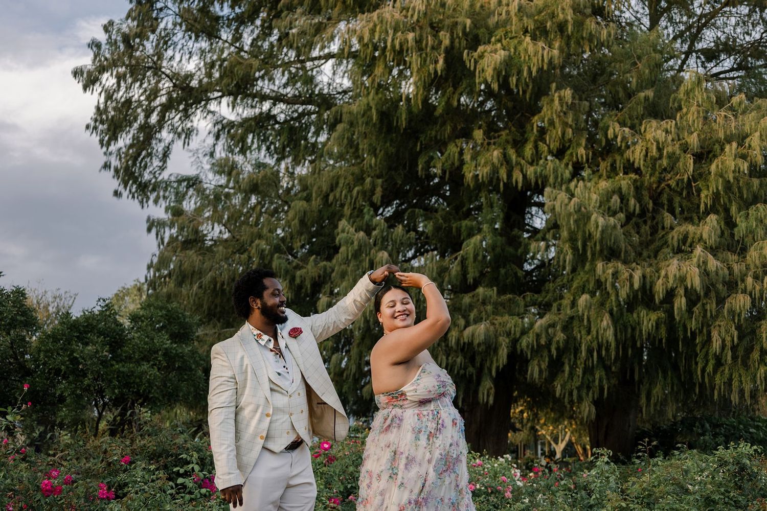 A joyful couple dances together in a garden setting with a willow tree and cloudy sky in the background.