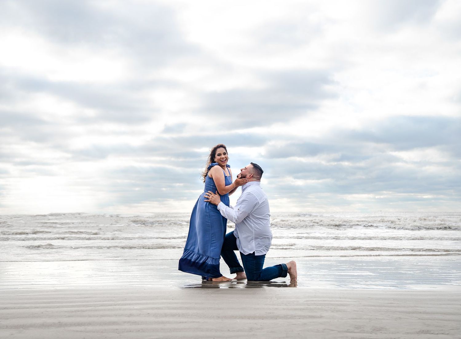 A romantic beach proposal scene with two people in blue and white outfits on a cloudy day by the ocean.