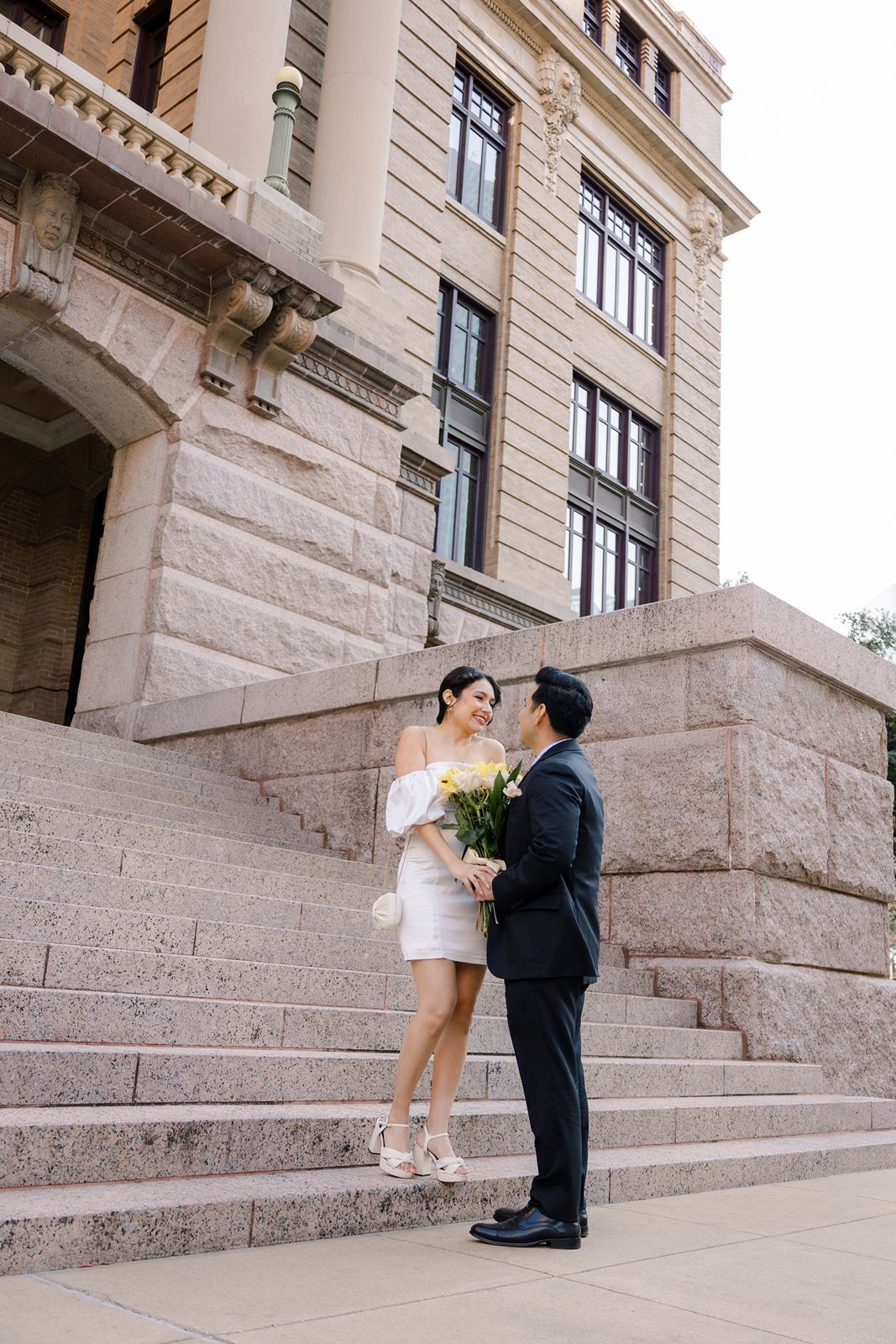 A couple in wedding attire stands on grand stone steps outside a historic building with cream-colored walls.
