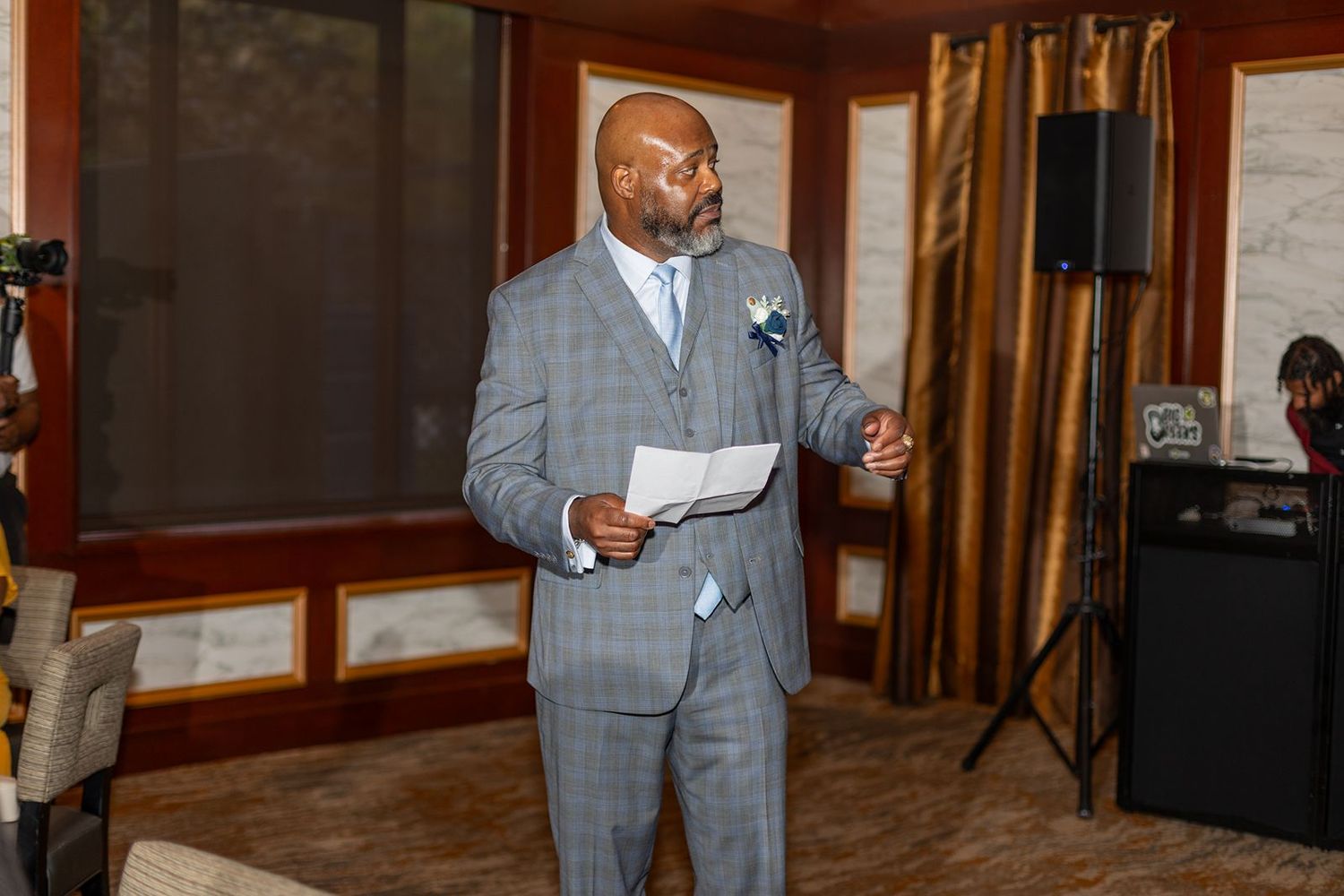 Man in gray plaid suit speaking at a wedding reception while holding papers.