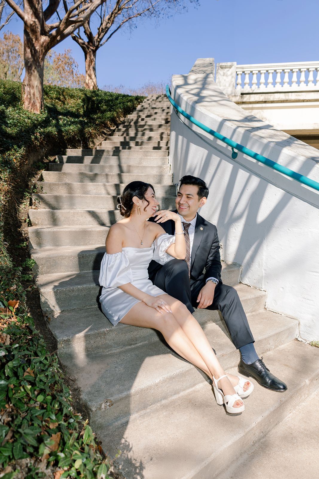 Couple in formal attire sitting together on outdoor stone steps, smiling at each other in a sunny park setting.