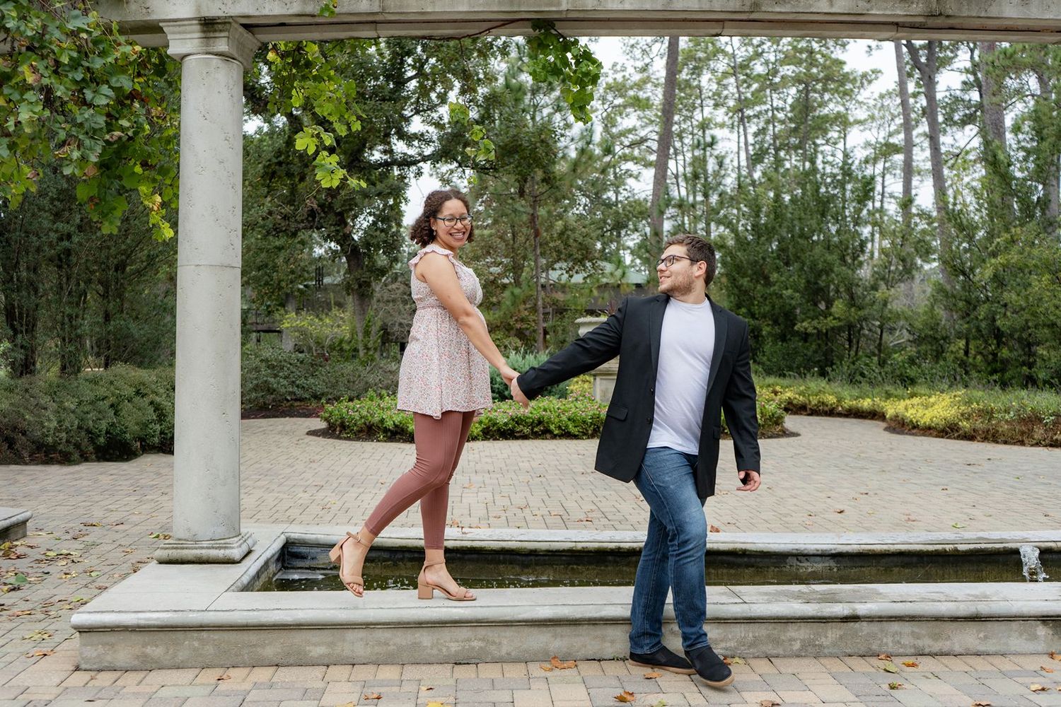 Couple holding hands under a garden pergola, woman in pink dress and man in blazer, smiling outdoors.